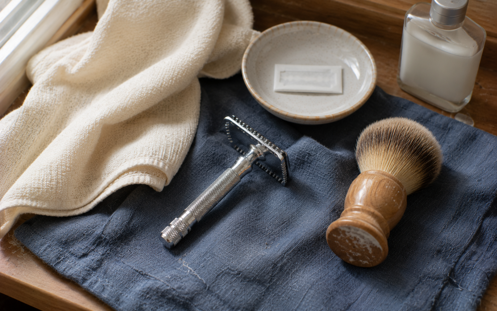 A safety razor and shaving brush resting on a folded warm cream cloth on slate blue linen in natural daylight