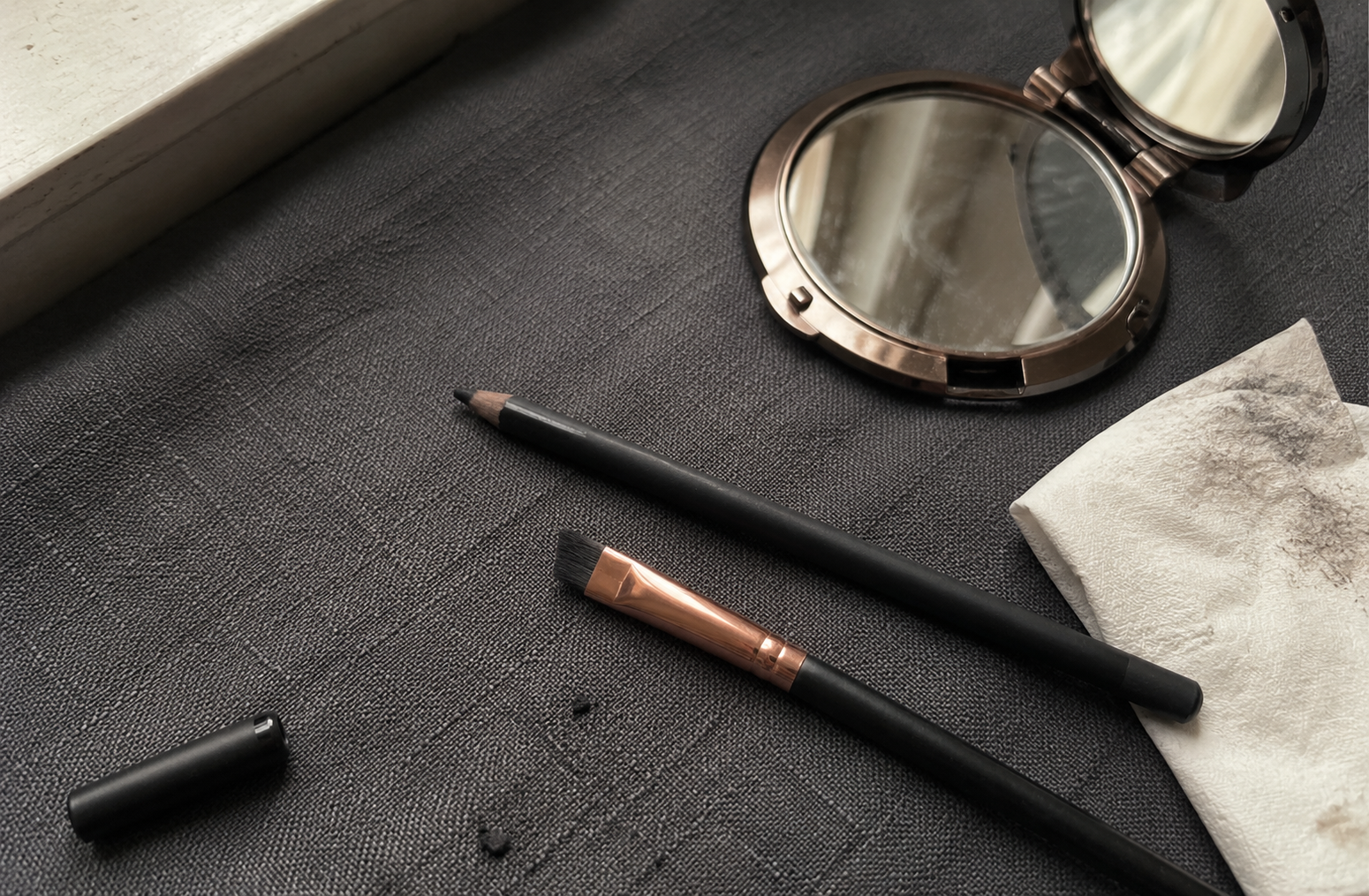 A black eyeliner pencil and small angled brush resting on a soft charcoal surface in gentle natural light