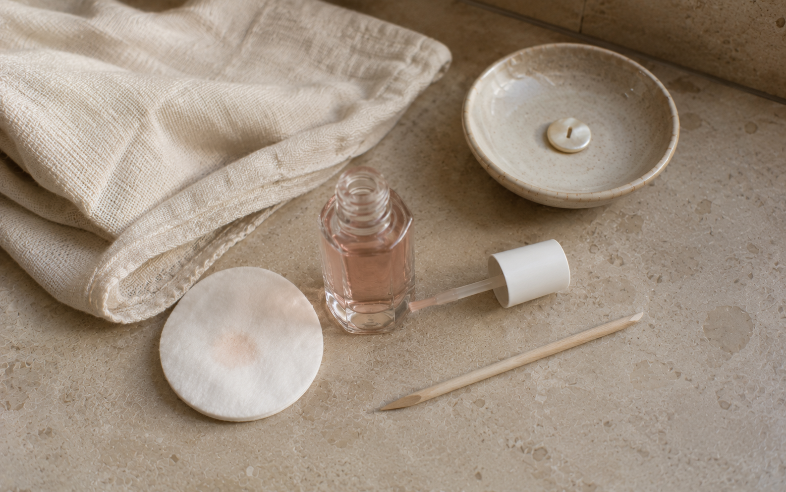 A small bottle of cuticle oil and a wooden manicure stick on soft pink-toned linen in natural daylight