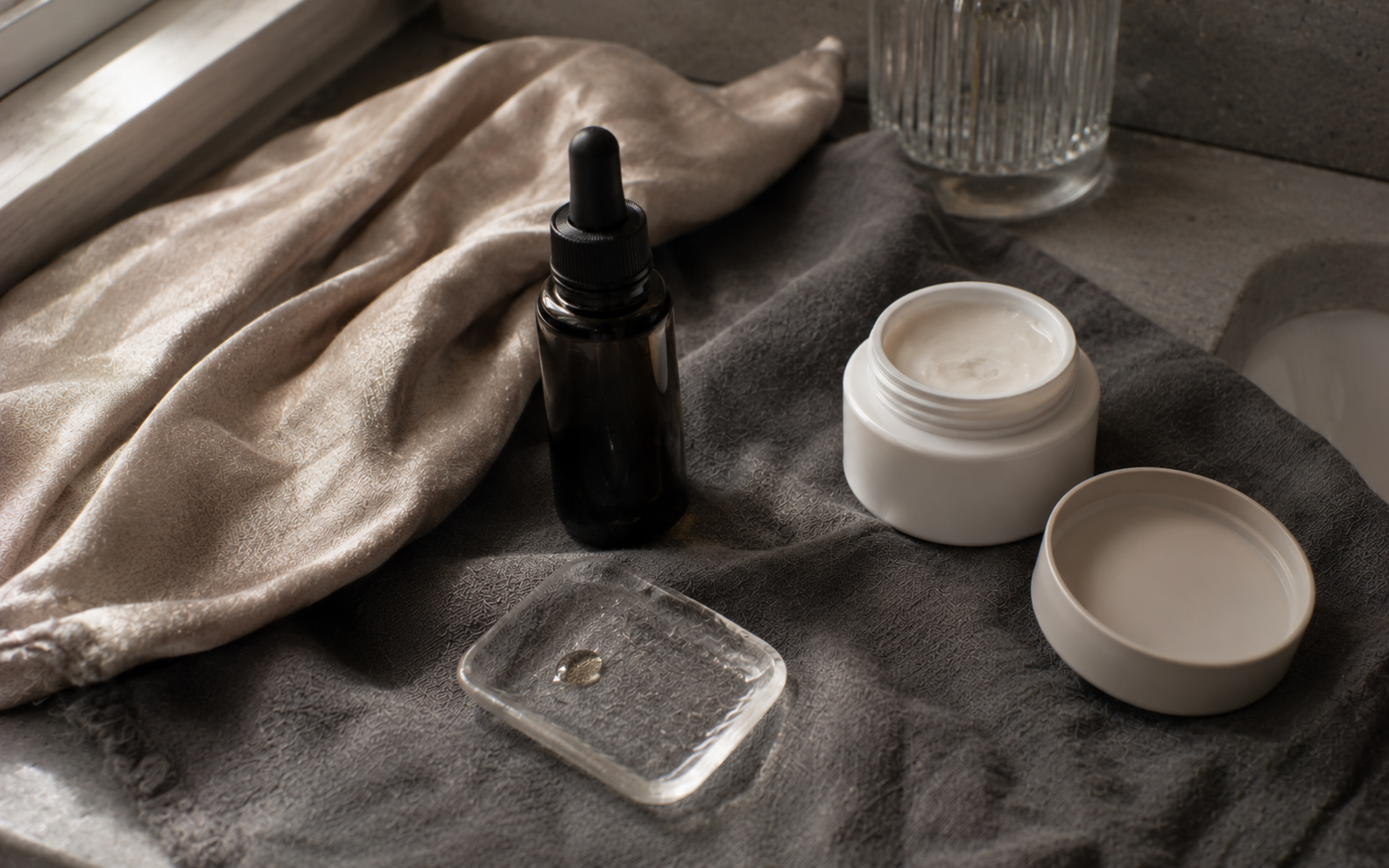 A small bottle of serum beside a folded soft champagne-toned cloth on smoky pewter linen in natural daylight