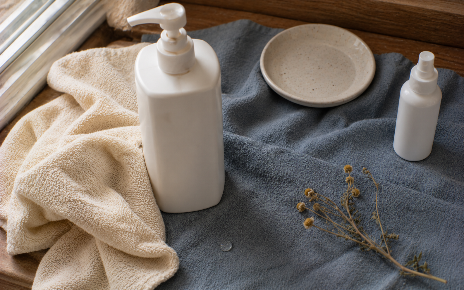 A bottle of body wash beside a soft chamomile-toned cloth on dusty steel blue linen in natural daylight