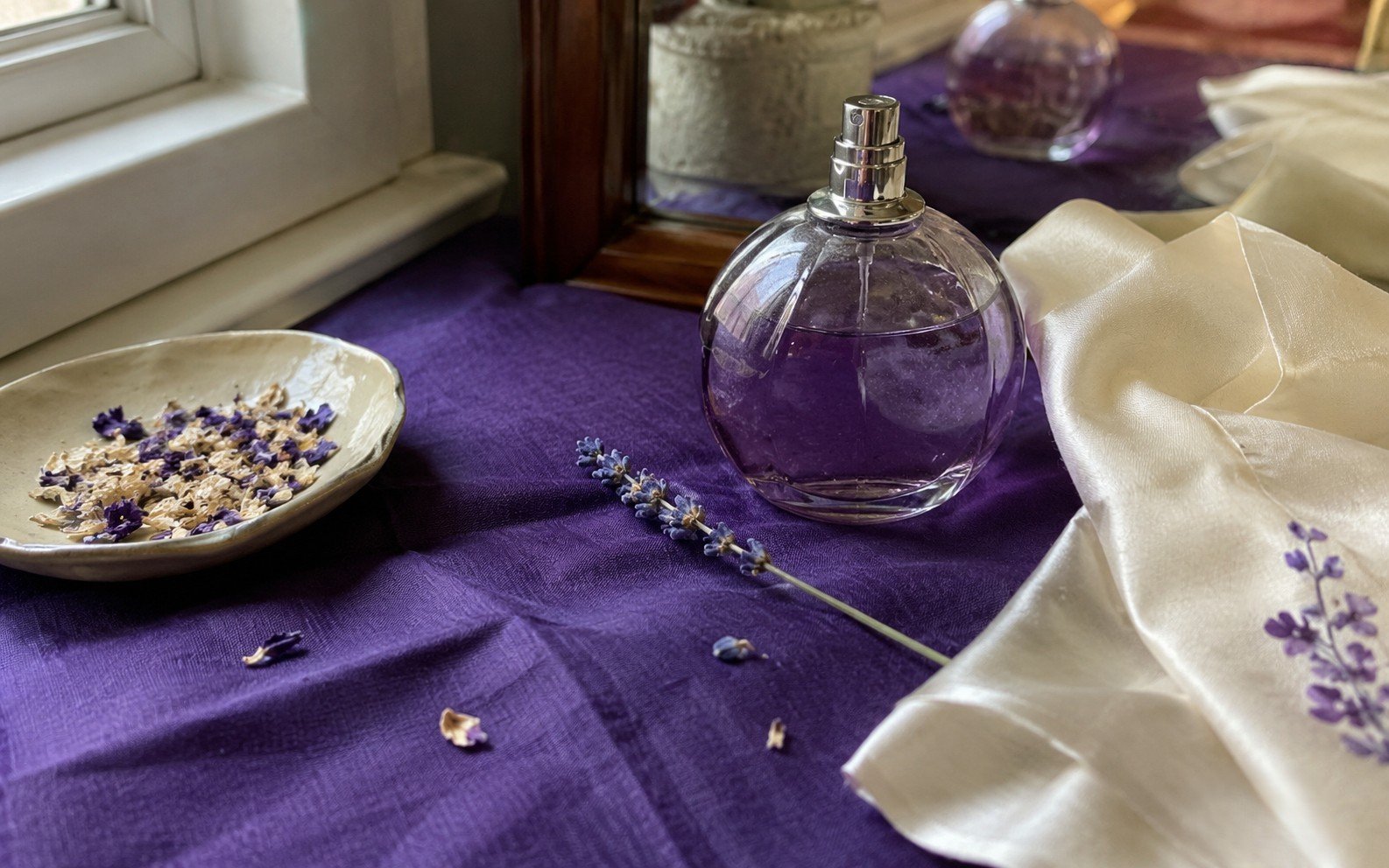 A small perfume bottle resting on rich violet linen with a single dried flower in soft natural light